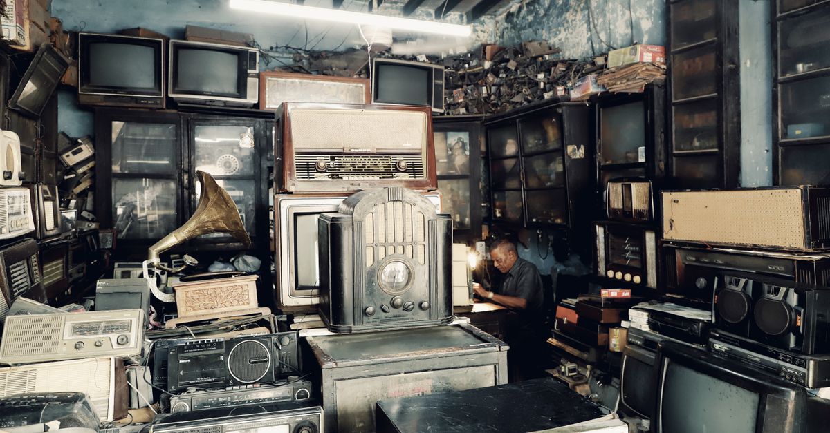 A cluttered vintage shop filled with antique radios, TVs, and a man working in the background.